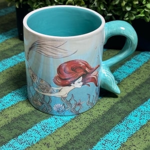 May include: A ceramic mug with a light blue exterior and interior. The mug features a graphic illustration of a mermaid with long red hair swimming among seaweed and shells. The handle is shaped like a mermaid tail in a matching light blue color.