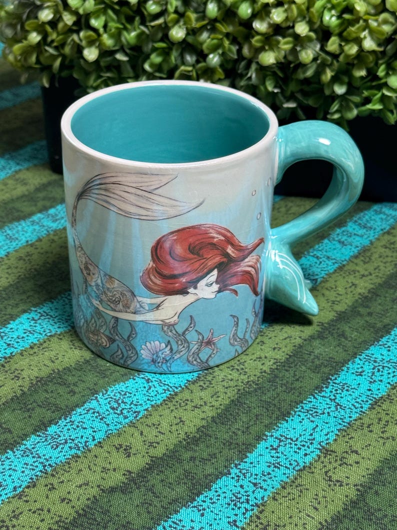 May include: A ceramic mug with a light blue exterior and interior. The mug features a graphic illustration of a mermaid with long red hair swimming among seaweed and shells. The handle is shaped like a mermaid tail in a matching light blue color.