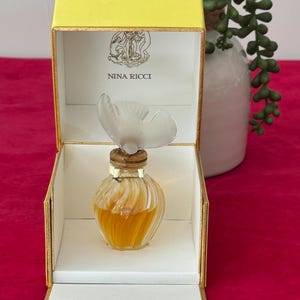 Op de afbeelding: Een vintage Nina Ricci parfumflesje in een gele en witte doos. De parfumfles heeft een doorzichtige, vlindervormige stopper. De doos is open en toont het parfumflesje en de woorden "NINA RICCI". De parfum binnenin is amberkleurig.