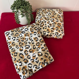 May include: Two folded fabric pieces with a leopard print pattern in shades of tan, brown, and black on a white background. The fabric is placed on a red surface, with a small potted plant in the background.