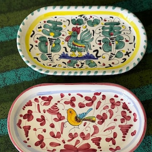 May include: Two oval ceramic serving platters. The top platter has a rooster design with green, yellow, and blue accents. The bottom platter features a yellow bird and red floral pattern with a red border. Both are hand-painted.