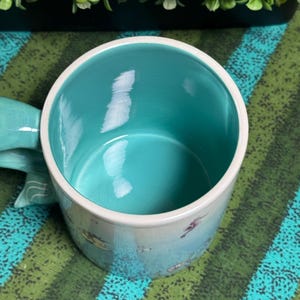 May include: A ceramic mug with a smooth, bright turquoise interior and a white rim. The exterior features a light blue base color with small, scattered illustrations of sea creatures. A curved handle extends from the side of the mug.