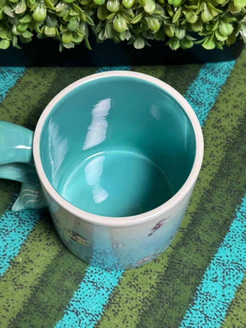 May include: A ceramic mug with a smooth, bright turquoise interior and a white rim. The exterior features a light blue base color with small, scattered illustrations of sea creatures. A curved handle extends from the side of the mug.