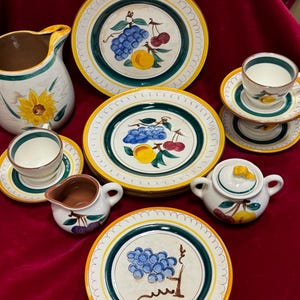 May include: A vintage tea set with a fruit motif. The set includes a pitcher, a sugar bowl, a creamer, two teacups, and two saucers. The pieces are decorated with a colorful design of grapes, cherries, and peaches.
