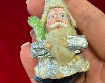 May include: Vintage Christmas ornament depicting Santa Claus. The ornament features a silver cone-shaped hat, a white beard, and a painted face. Santa is holding a green tree. The ornament is made of fabric and has a string for hanging.