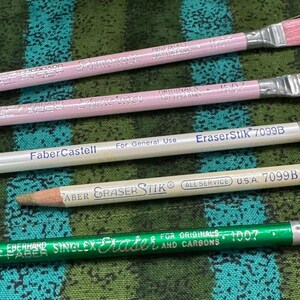 May include: A selection of pencils and erasers, including pink-handled paint brushes, a white Faber-Castell pencil, and a green Eberhard Faber eraser. The items are displayed on a patterned green and blue fabric.