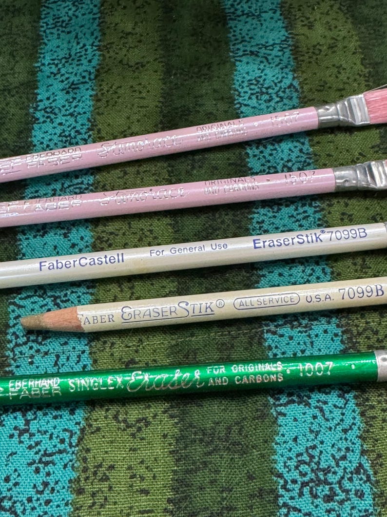 May include: A selection of pencils and erasers, including pink-handled paint brushes, a white Faber-Castell pencil, and a green Eberhard Faber eraser. The items are displayed on a patterned green and blue fabric.
