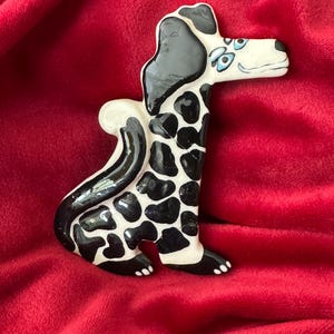 May include: A ceramic dog figurine with a black and white spotted pattern. The dog has a white face with blue eyes and a black ear. The figurine is sitting on a red surface.