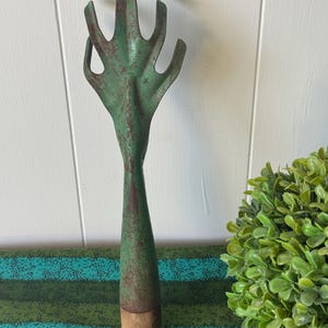 May include: A vintage garden hand rake with a green, weathered finish. The rake head has five tines, and the handle is a tapered shape with a wooden base. The tool shows signs of age and use, with rust visible on the metal.