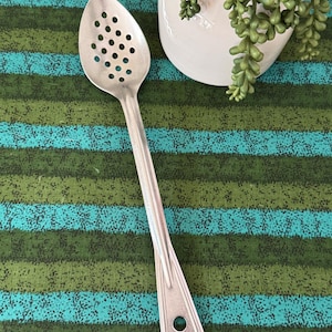 May include: A silver-toned metal slotted spoon featuring a perforated oval bowl and a long, slender handle with a circular hole at the end for hanging. The spoon has a smooth, metallic finish and a simple, functional design for kitchen use.