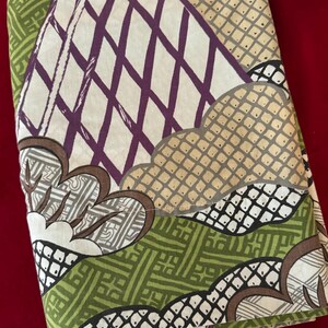 May include: Folded fabric with a repeating pattern in shades of green, purple, tan, and white. The design includes geometric shapes, diamond patterns, and stylized floral elements. The fabric is folded, showing multiple layers.