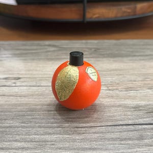 May include: A small, round, orange bottle with a black cap. A gold leaf design is on the side. The bottle has a small label that reads "Lauderdale Florida".