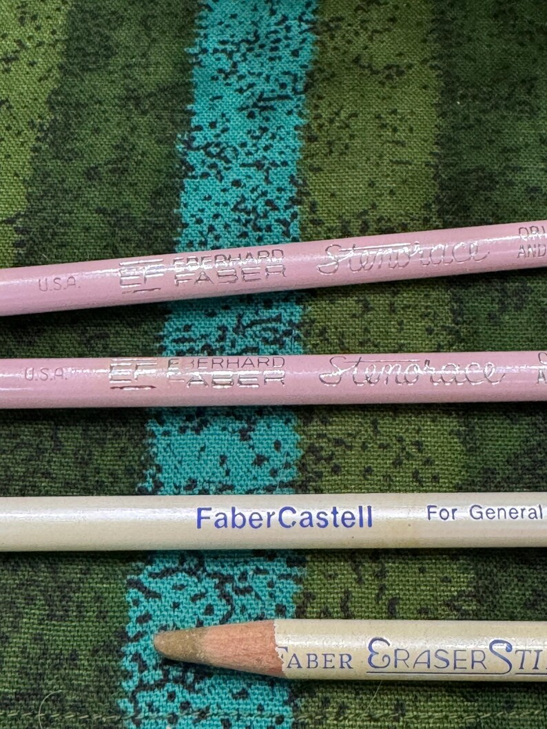 May include: Close-up of four pencils. Two are light pink with "Eberhard Faber" and "Stenotrace" printed on them. One is white with "Faber-Castell" printed on it. The last is white with "FABER ERASER STICK" printed on it.