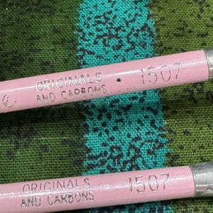 May include: Close-up of two pink pencils with silver metal ends. The pencils have the text "ORIGINALS AND CARBONS" and the number "1507" printed on them. The pencils are set against a green and teal patterned fabric.