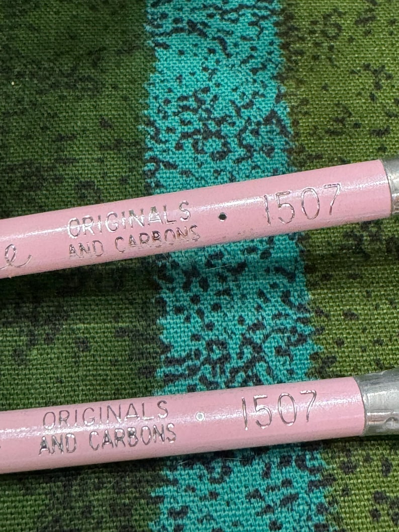 May include: Close-up of two pink pencils with silver metal ends. The pencils have the text "ORIGINALS AND CARBONS" and the number "1507" printed on them. The pencils are set against a green and teal patterned fabric.