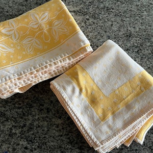 May include: Two folded yellow and white patterned tablecloths. One tablecloth has a floral design border, and the other has a geometric border. The tablecloths are made of a soft fabric and are folded neatly.