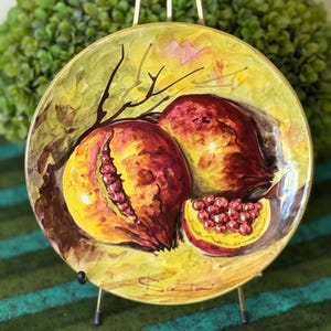 May include: A decorative ceramic plate featuring a hand-painted design of pomegranates. The plate has a yellow and green background with a brown border and a gold stand. The pomegranates are red and yellow with a detailed depiction of the seeds.
