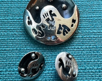 May include: A silver-toned jewellery set featuring a large round brooch and two oval earrings. The brooch and earrings have a yin-yang design with a stylized bird or animal figure. The set is displayed on a teal textured surface.