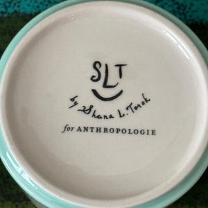 May include: A ceramic bowl with a light blue rim and a white interior. The bottom of the bowl features the text "SLT" above a smiling face, followed by "by Shana L. Torok" and "for ANTHROPOLOGIE" in black lettering.