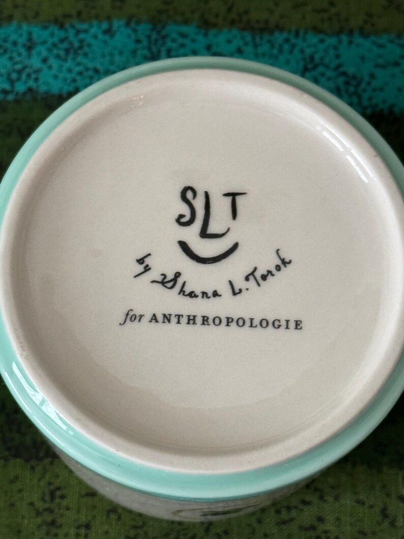 May include: A ceramic bowl with a light blue rim and a white interior. The bottom of the bowl features the text "SLT" above a smiling face, followed by "by Shana L. Torok" and "for ANTHROPOLOGIE" in black lettering.