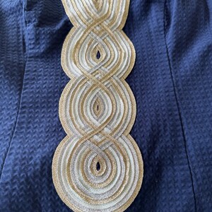 Pu&ograve; includere: Primo piano di un capo blu navy con un motivo strutturato. La scollatura &egrave; adornata da un intricato design di finiture intrecciate oro, argento e bianco, creando un motivo circolare a strati. Il tessuto sembra essere un materiale tessuto.