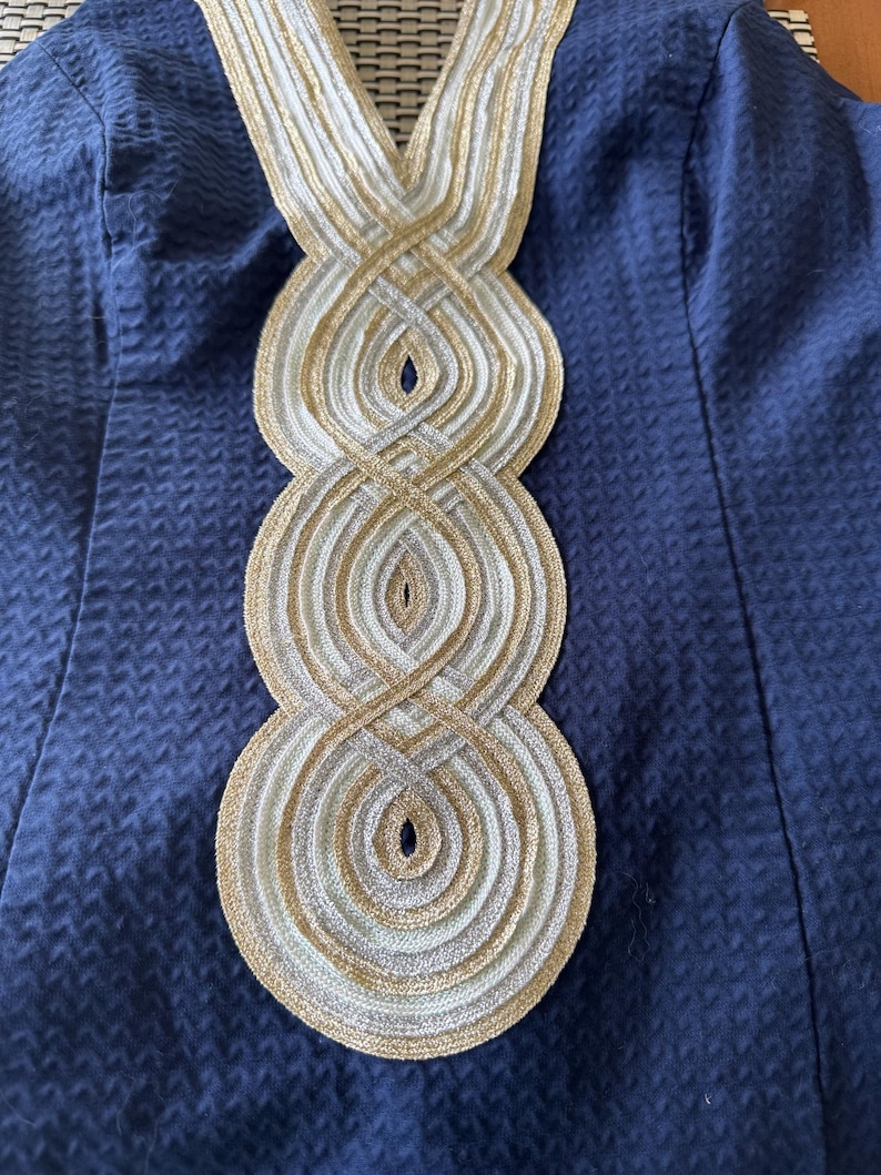 Pu&ograve; includere: Primo piano di un capo blu navy con un motivo strutturato. La scollatura &egrave; adornata da un intricato design di finiture intrecciate oro, argento e bianco, creando un motivo circolare a strati. Il tessuto sembra essere un materiale tessuto.