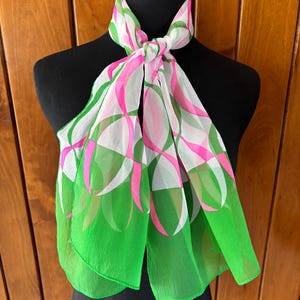 May include: A sheer scarf with a vibrant floral design. The scarf features a pattern of pink and white overlapping shapes against a green and white background. The scarf is draped over a black mannequin, showcasing its flowing fabric.