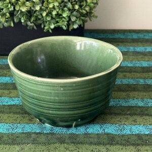 May include: A round ceramic bowl with a glossy sage green glaze and a subtle crackle finish. The exterior of the bowl features a series of horizontal, raised ridges that wrap around the circumference, creating a textured, tiered appearance.