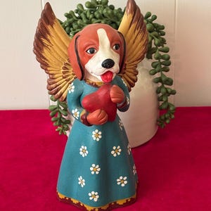 May include: A ceramic dog angel figurine with brown and gold wings, wearing a teal dress with white daisy accents, and holding a red heart. The dog has a white and brown face and a red tongue. The figurine is on a red surface.