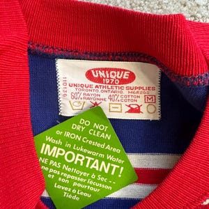 May include: A blue and white striped shirt with a red collar and red trim. The shirt has a label that reads "Unique 1970 Unique Athletic Supplies Toronto, Ontario, Mer2GS 60% Rayon 40% Cotton" and instructions for washing and ironing the shirt.