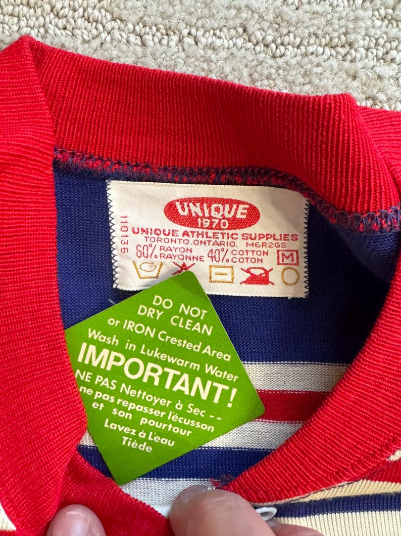 May include: A blue and white striped shirt with a red collar and red trim. The shirt has a label that reads "Unique 1970 Unique Athletic Supplies Toronto, Ontario, Mer2GS 60% Rayon 40% Cotton" and instructions for washing and ironing the shirt.