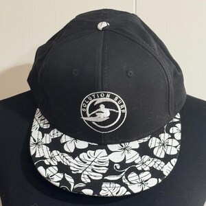 May include: Black baseball cap with a white and black floral patterned brim. The cap features the text "SOLUTION SURF" in a white circular logo with a surfer graphic.