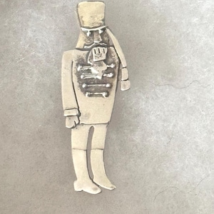 Pode incluir: Uma estatueta de metal prateado de um soldado quebra-nozes. A estatueta está em pé, usando um chapéu alto, um casaco com botões decorativos e botas. O design é detalhado com linhas e características gravadas.