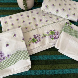 May include: A white bed sheet set featuring a repeating pattern of small purple violet flowers. The edges are decorated with a wider band of larger violet clusters and a green cross-hatched lattice border. The set includes a flat sheet and pillowcases.