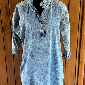May include: Acid wash denim dress with a mandarin collar and long sleeves. The dress features a button-down placket and a relaxed fit. The sleeves are cuffed at the elbow. The dress is displayed on a black mannequin.
