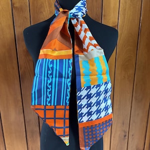 May include: A colorful silk scarf with a variety of geometric patterns. The scarf features orange, blue, and white designs, including stripes, houndstooth, and grid patterns. The scarf is displayed on a black mannequin.