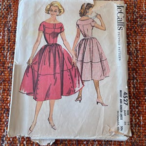 May include: Vintage McCall's printed pattern for a misses' and junior dress, with illustrations of the dress design. The pattern shows a red dress with a fitted bodice and a full skirt, and a pink checkered dress. The pattern is size 14, bust 86 cm.