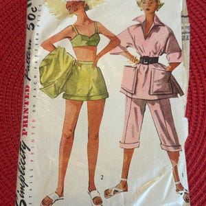 May include: Vintage Simplicity sewing pattern for a two-piece outfit. The pattern is for size 11, bust 29 inches. The pattern shows a woman wearing a green and white swimsuit top and shorts, and another woman wearing a pink shirt and shorts.