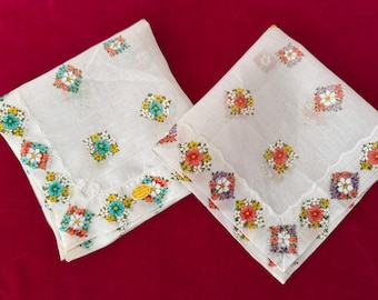 May include: Two white handkerchiefs with colorful floral embroidery. Each handkerchief features a scalloped edge and square floral patterns in red, yellow, blue, and white. A small gold tag is visible on one.