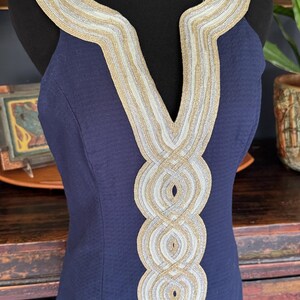 Pu&ograve; includere: Un abito blu navy con finiture decorative oro e argento intorno allo scollo e al corpetto. L'abito &egrave; senza maniche e ha un tessuto strutturato. La finitura presenta un motivo intrecciato e aggiunge un tocco di eleganza all'indumento.
