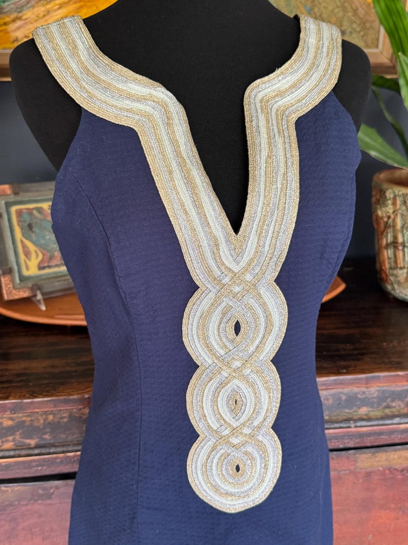 Pu&ograve; includere: Un abito blu navy con finiture decorative oro e argento intorno allo scollo e al corpetto. L'abito &egrave; senza maniche e ha un tessuto strutturato. La finitura presenta un motivo intrecciato e aggiunge un tocco di eleganza all'indumento.