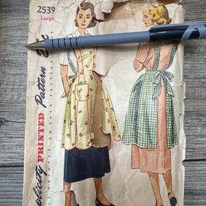 Op de afbeelding: Vintage Simplicity patroon 2539, maat Large, met illustraties van schorten. Het patroon staat op verouderd papier, met een blauwe pen eroverheen. De schorten hebben bloemen- en ruitmotieven.