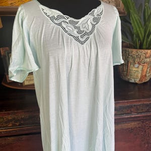May include: A light blue nightgown featuring short sleeves and a V-neckline adorned with white floral lace embroidery. The garment has a loose, flowing silhouette with subtle gathers at the neckline, providing a simple and comfortable design.