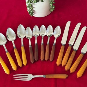 May include: A collection of vintage silverware, including spoons, knives, and a fork, arranged on a red surface. The spoons and knives have silver blades and handles with a mix of amber and brown tones. The fork has a matching amber handle.