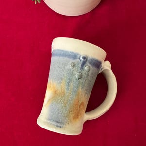 May include: Handmade ceramic mug with a white handle and rim, featuring a blue and yellow speckled design. The mug has three small, raised circles on the front and is set against a red background. A white ceramic bowl and greenery are in the background.