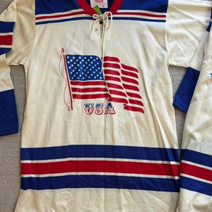 May include: A white hockey jersey with blue and red stripes. The jersey features a large American flag graphic with the word "USA" printed below it.
