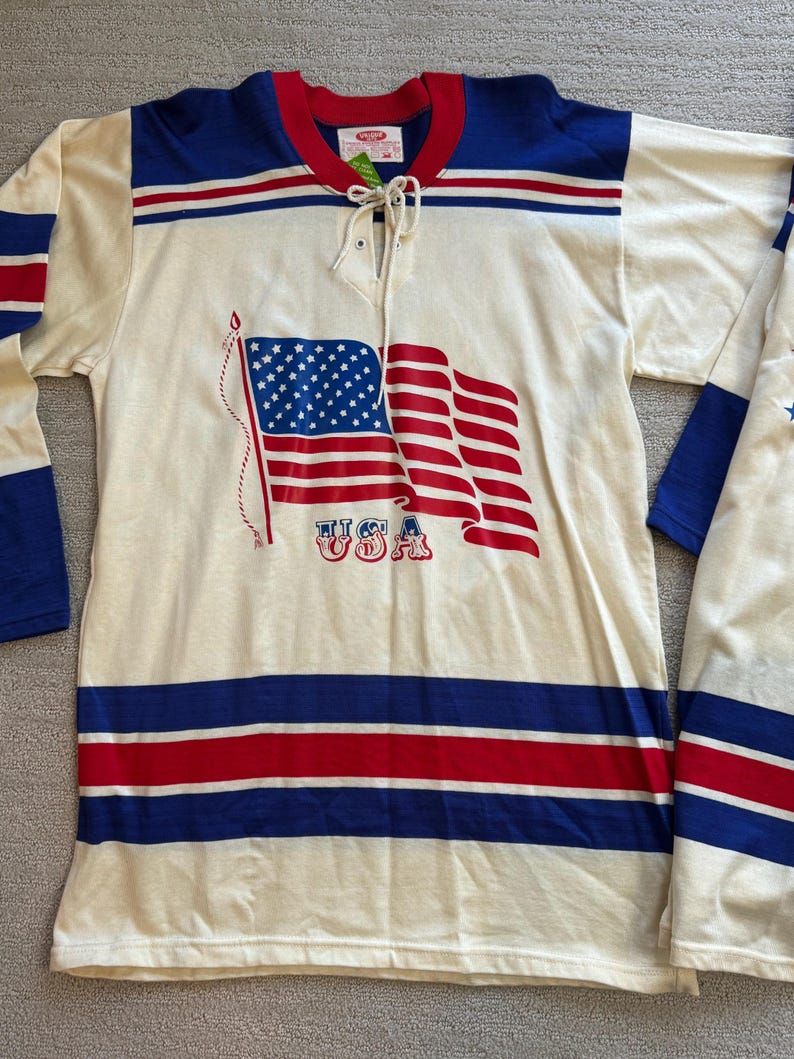 May include: A white hockey jersey with blue and red stripes. The jersey features a large American flag graphic with the word "USA" printed below it.