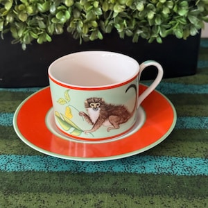 May include: A porcelain teacup and saucer set featuring a light green background with a detailed illustration of a brown monkey holding a yellow fruit on a branch. The rim of the cup and the border of the saucer are accented with a vibrant orange band.