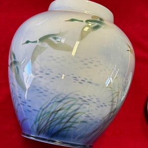 May include: A white ceramic vase with a blue and green glaze. The vase features a design of ducks flying over a body of water with reeds in the foreground.