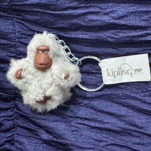 Peut inclure: Un porte-clés singe en peluche blanche avec des traits du visage et des pieds bruns. Le porte-clés comprend une chaîne argentée et un anneau métallique. Une étiquette blanche avec la marque "Kipling" et un logo de singe est attachée.
